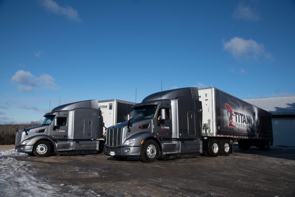 Two Titan Transline transport trucks parked outside a warehouse with branded trailers under a blue sky.