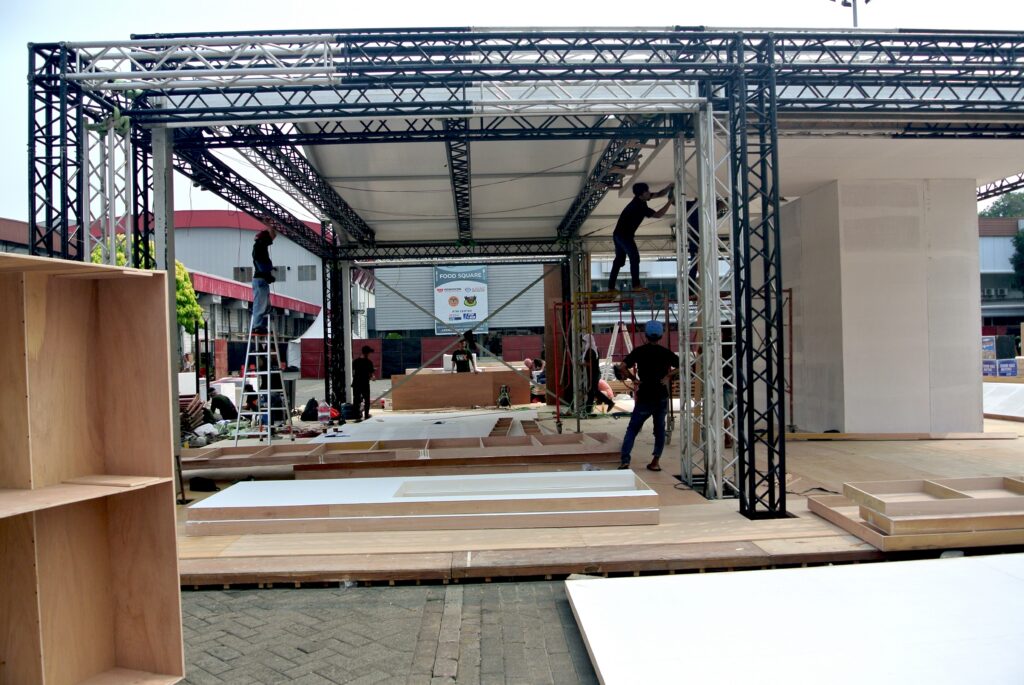 Workers assembling a large tradeshow booth structure with staging materials, ladders, and display panels.